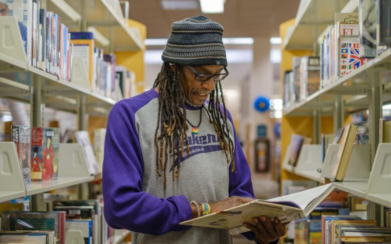 Brian Bukle wears a gray beanie cap and a t-shirt with purple sleeves, and reads a book in the aisle of the public library.