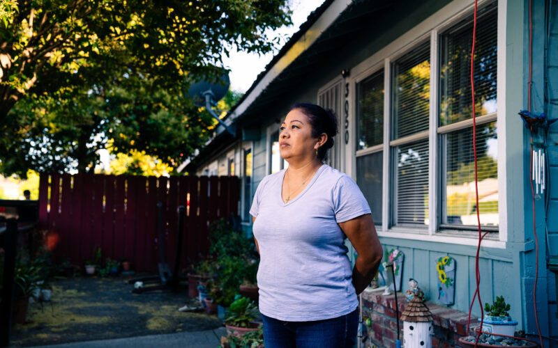 Maricela stands in front of her home, looks into the distance with her hands behind her back.