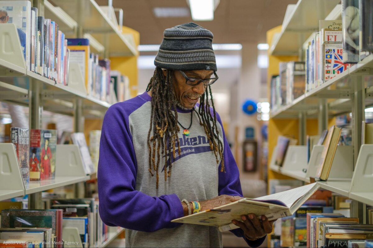 Brian Bukle wears a gray beanie cap and a t-shirt with purple sleeves, and reads a book in the aisle of the public library.