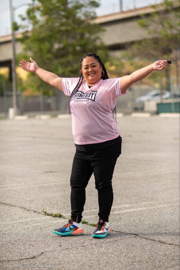Maria Luna stands in a parking lot, smiling with her arms open. She is wearing a pink jersey shirt and black pants, and colorful sneakers.