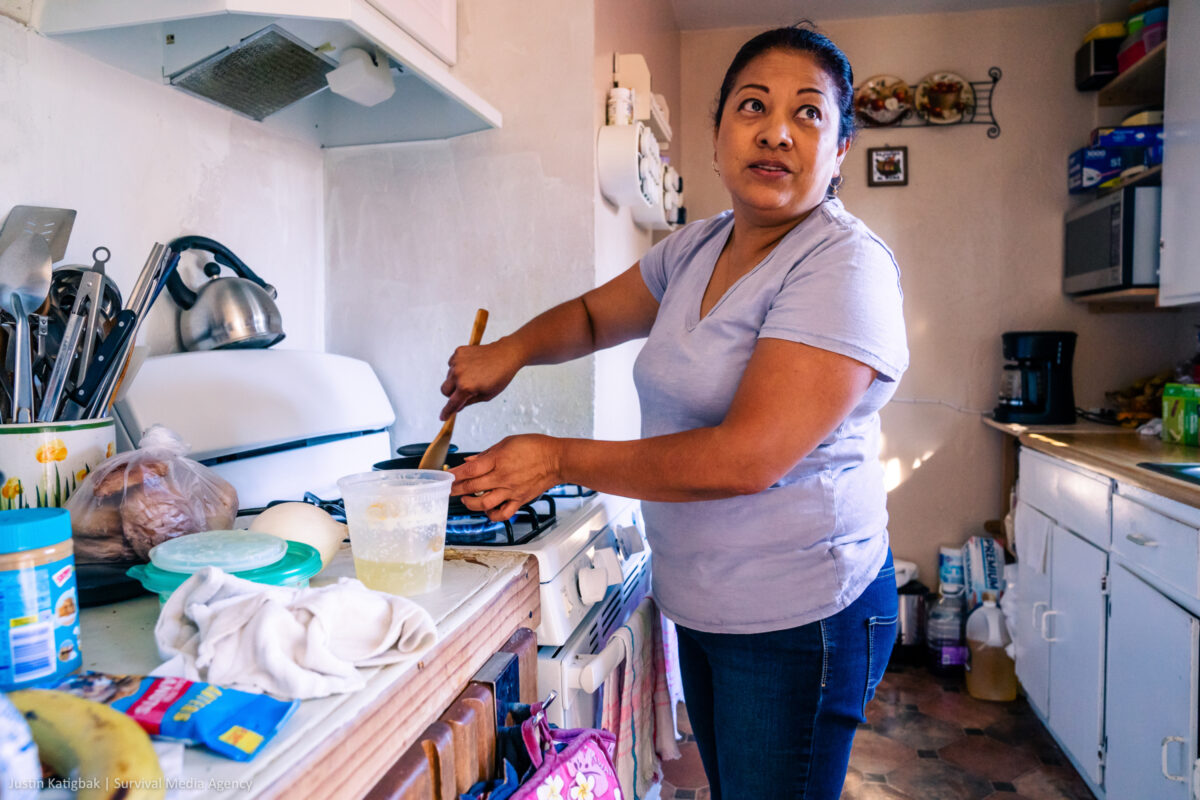 Maricela stands in her kitchen and stirs a pot of food, smiling back.