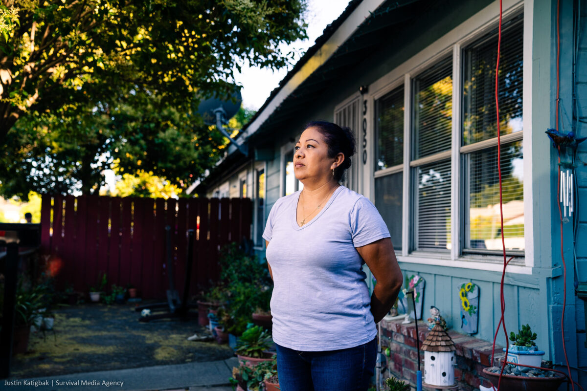 Maricela stands in front of her home, looks into the distance with her hands behind her back.