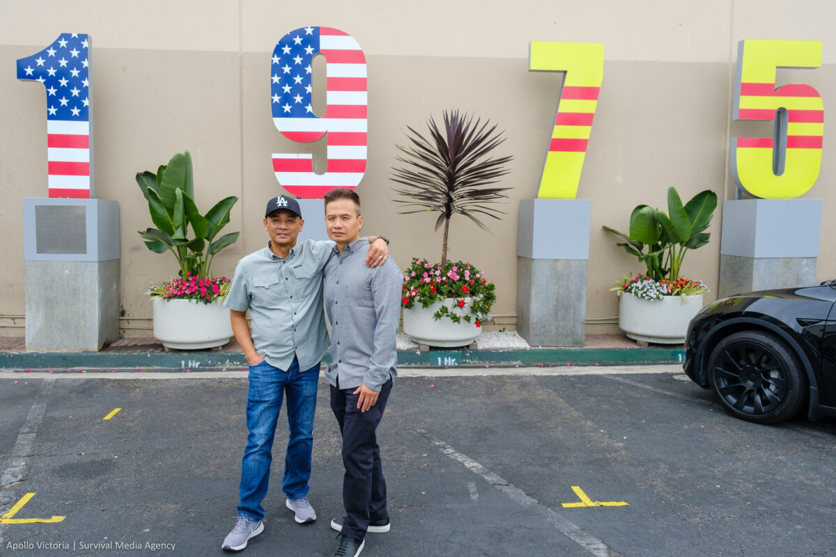 An wraps an arm across Tin's shoulder as they stand in front of four pillars with large numbers on top of them. Together, the numbers say, "1975" with half decorated with the stars and stripes of the U.S. flag and half decorated with the colors of the former South Vietnam flag.