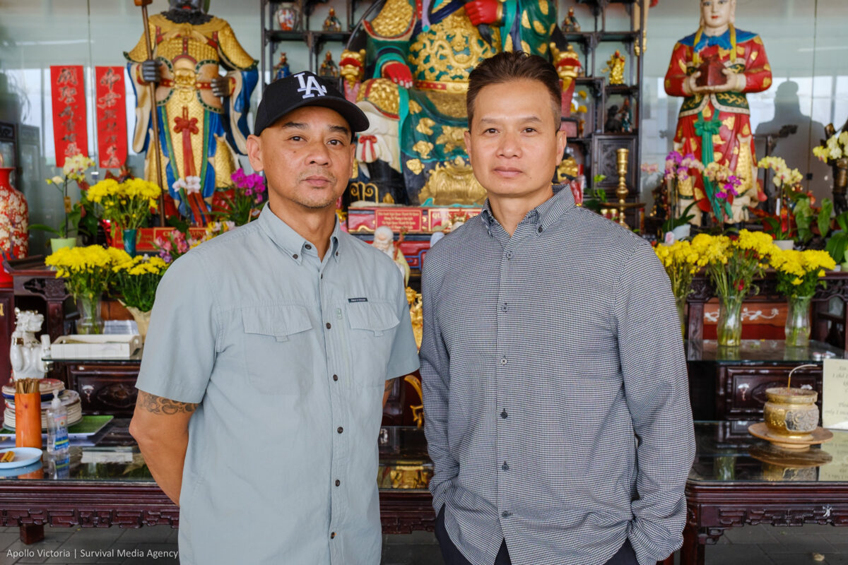 An and Tin pose for a photo in front of a large wooden altar laden with Buddhist figures, flowers, and incense. THey both wear button up shirts and look somber.