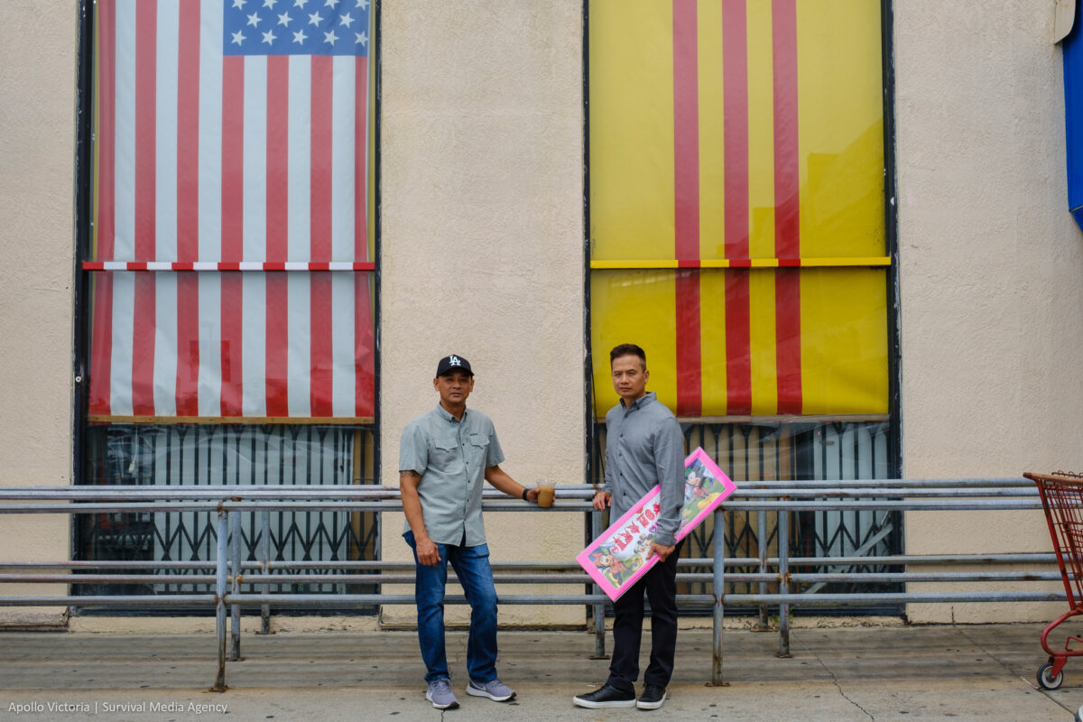 An and Tin stand in front of two large windows of the Asian Garden Mall building. One window has the U.S. flag hanging vertically and the South Vietnam flag hangs in the other.
