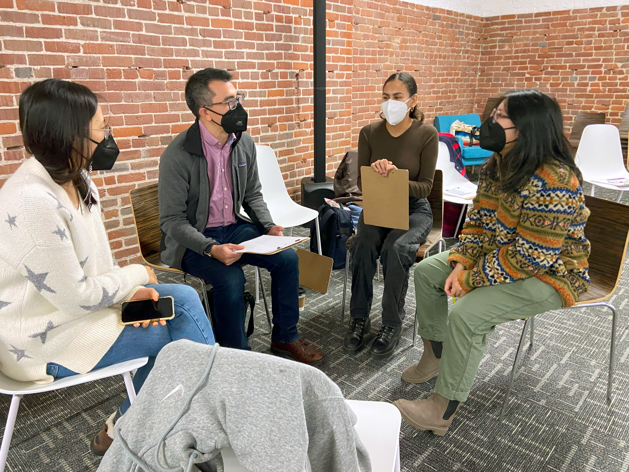 A group of people wear face masks and sit in a circle as they have a discussion. Some hold clipboards.