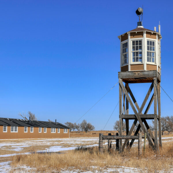 An octagonal wooden structure sits on stilts, paned windows spanning the circumference of the watch tower. Dry grass and ice stretch from the foreground to the background where a long, one-story building with several windows stands not too far from the watch tower. Much further away stands a water tower.
