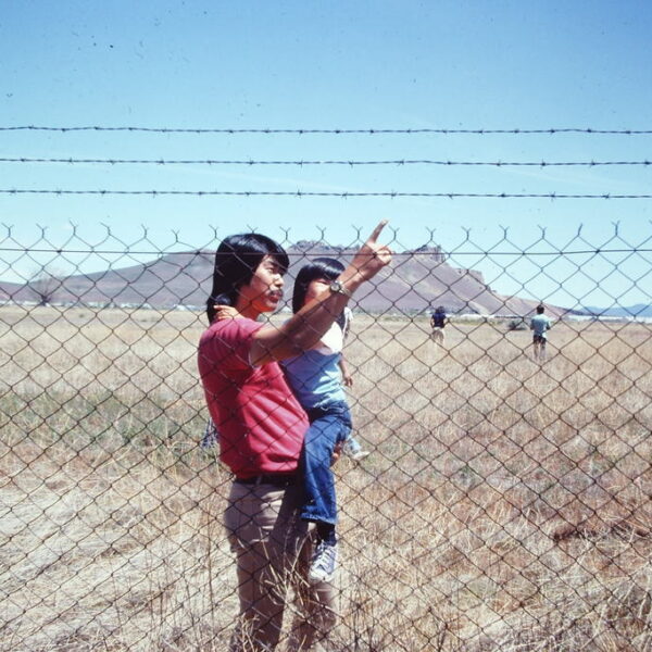 A man holds a child in one arm who looks as he touches the barbed wire fence in front of them. Dry grassland surrounds them. A little further behind is a group of other Japanese Americans participating in the Tule Lake pilgrimage. The hill called Castle Rock is in the background.