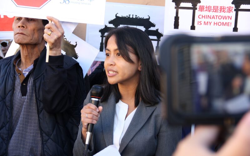 A woman in a blazer holds a microphone in front of people holding signs. One sign reads "Chinatown is my home"