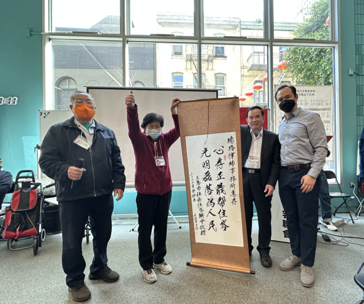 4 individuals pose together for a photo next to a scroll of Chinese calligraphy.