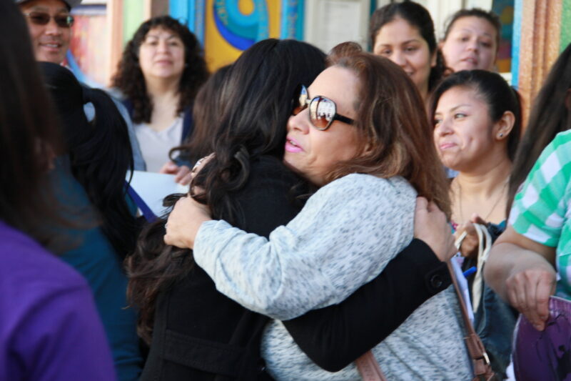 Two women are hugging in front of a group of people and one is wearing sunglasses