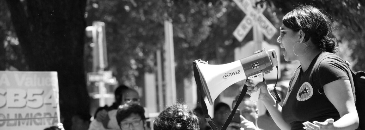 Black and white photo of a woman in a t-shirt holding a megaphone standing above a crowd of people in a park. In the background a person holds up a sign that says 'CA Values SB 54'
