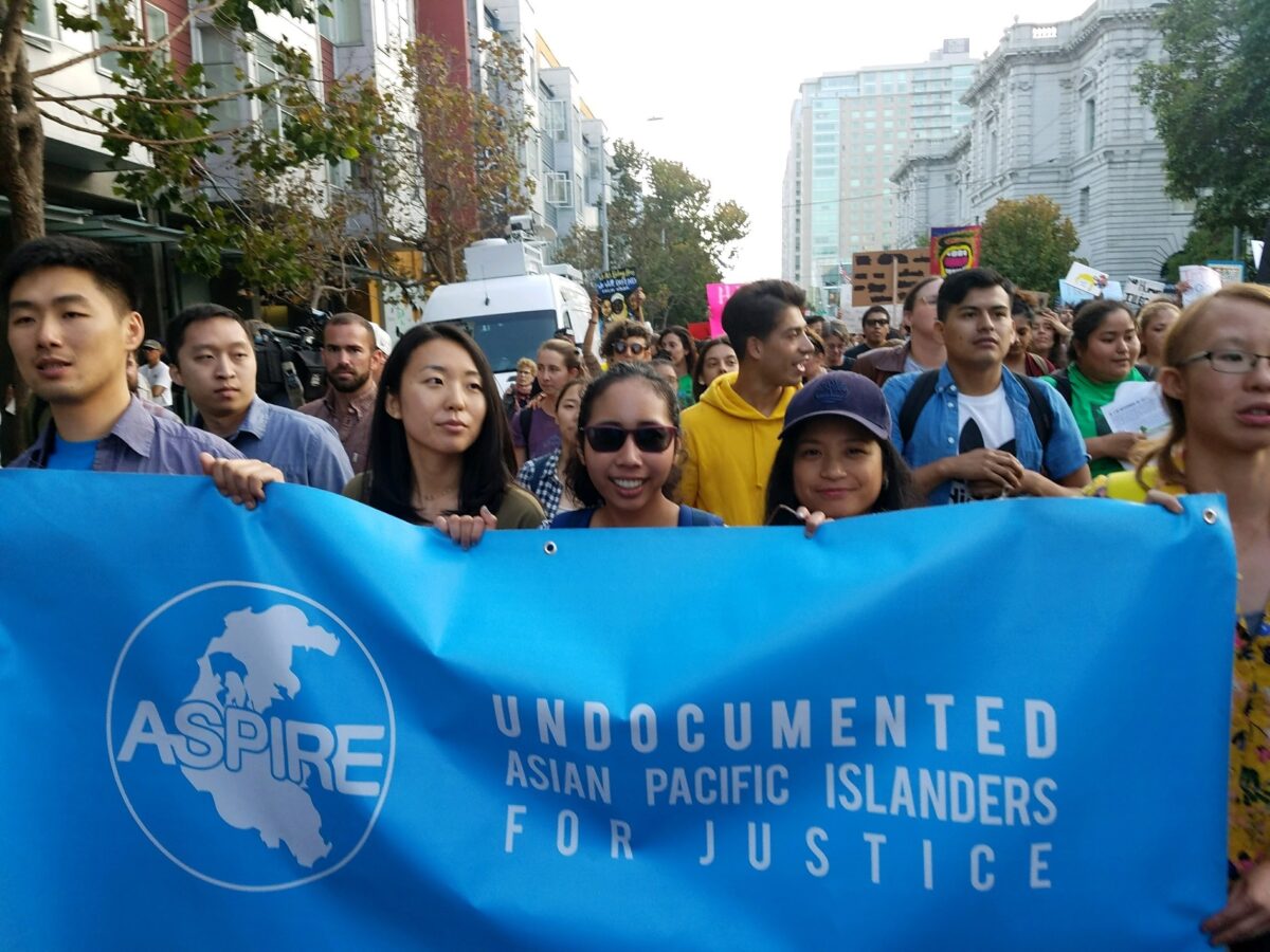 So Young stands in the middle of a row of five protestors walking down a city street, holding up a blue banner that reads: Undocumented Asian Pacific Islanders for Justice."