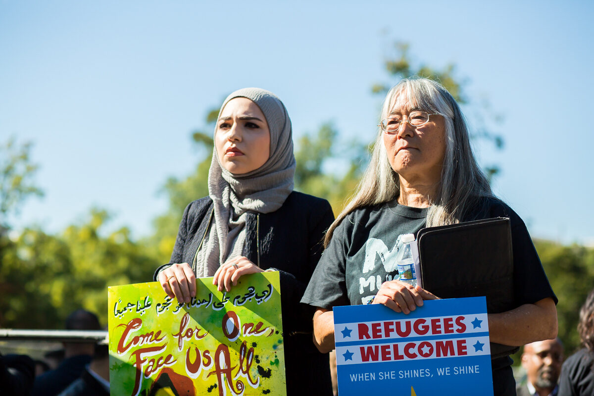 Two community members stand close together and hold signs that say "Refugees Welcome" and "Come for One, Face Us All."
