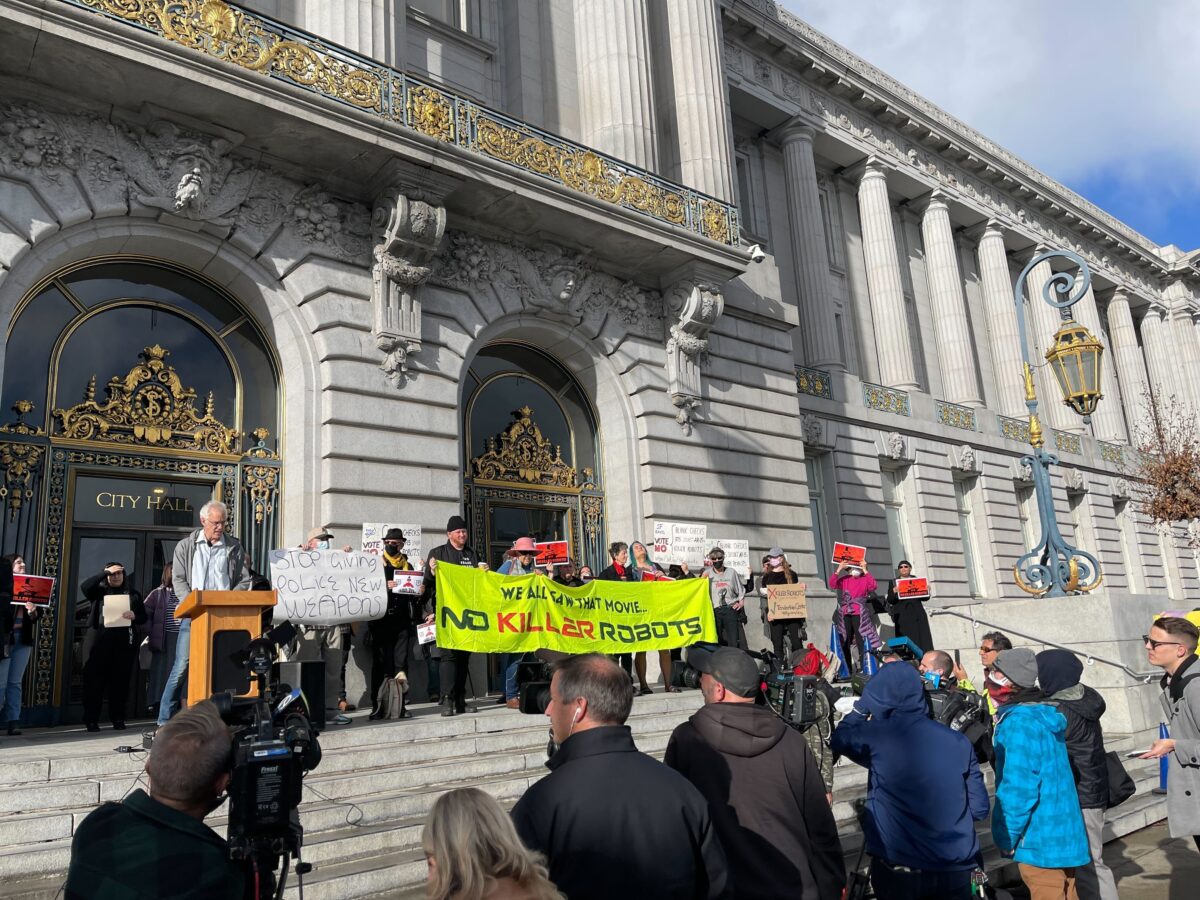 A crowd gathers at the foot of stone steps leading to a landing in front of San Francisco City Hall, an ornate building with neoclassical architectural features and gold-colored metal work. At the top of the stairs, in front of large doors, people stand beside a man in a suit speaking from a podum. Some of those standing hold a long banner that says, "We all saw that movie... No Killer Robots."