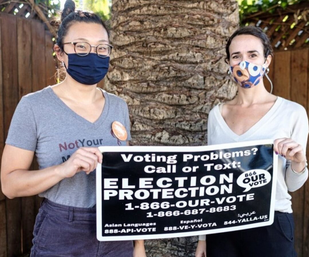 Two women wearing masks stand in front of a palm tree holding a sign with election protection information