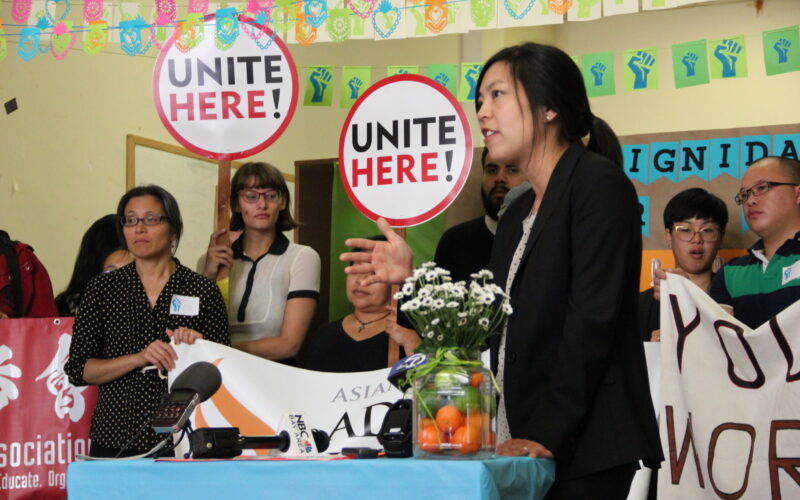 ALC attorney WInnie Kao speaks at a press conference behind a table with microphones and a vase with oranges and flowers. Behind her colorful bunting hangs from the ceilings and people hold banners and signs saying "Unite Here!"