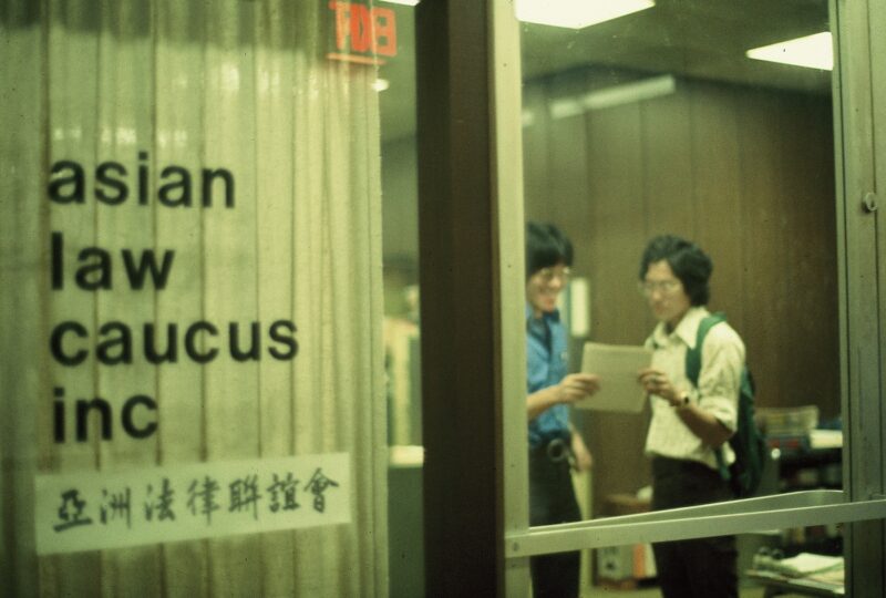 Office window reads "asian law caucus inc." on one side as conversation between two people happens behind left side of window