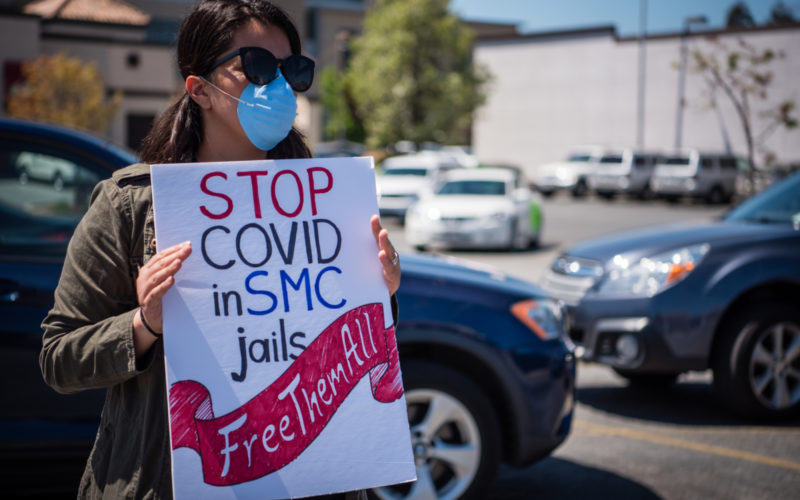 A woman in a blue surgical mask and sunglasses holds a sign reading "Stop COVID at SMC jails. Free them all"